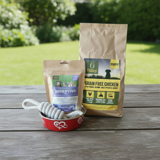 A bag of Hilltop Grain Free Chicken with Turkey & Salmon dog food for puppies and a pouch of Hilltop Probiotic Digestive treats are displayed on a wooden table next to a red bowl with a rope toy.
