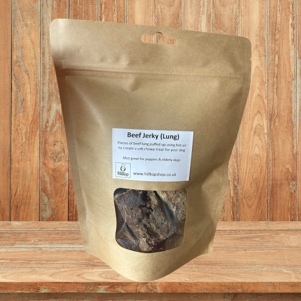A brown resealable bag of Hilltop Pet Foods Puffed Beef Jerky (Lung) sits on a wooden surface and background.